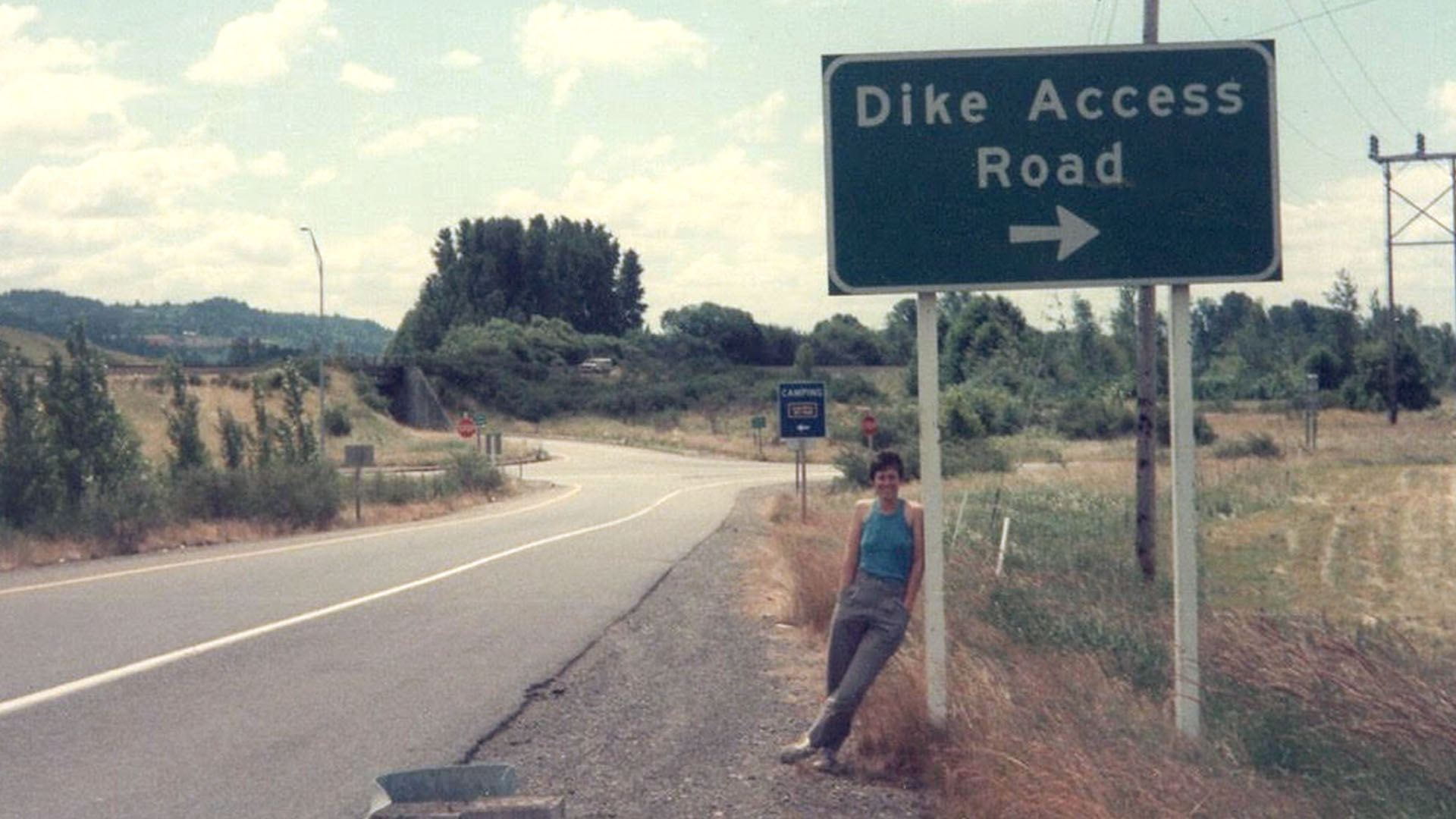 Judith Raiskin standing leans against Dike Access Road highway guide sign in Oregon.